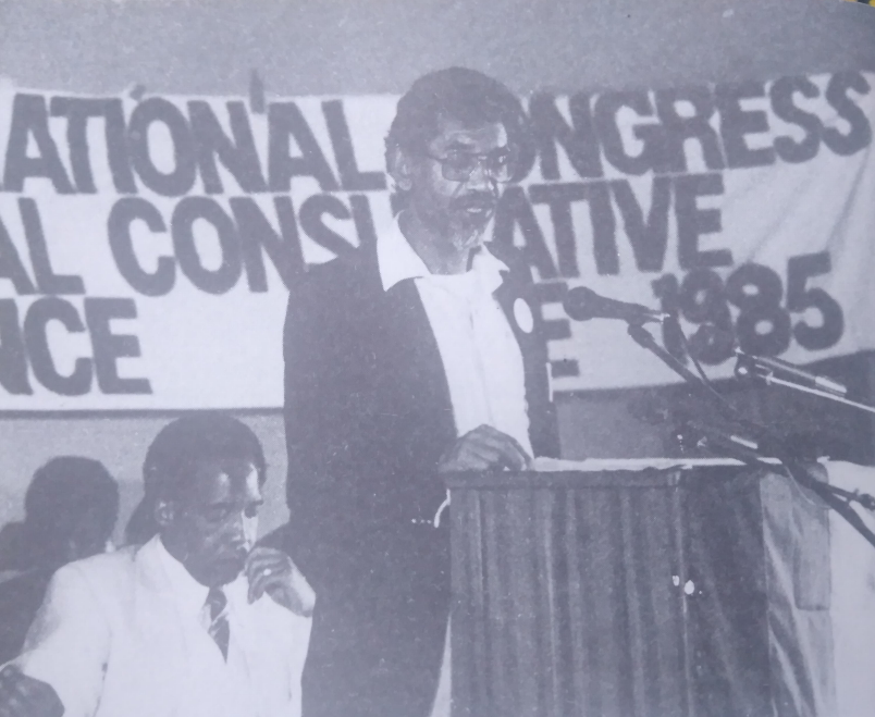 Photo of Mac Maharaj speaking at the Kabwe Conference, 1985. Seated behind him is Chris Hani.